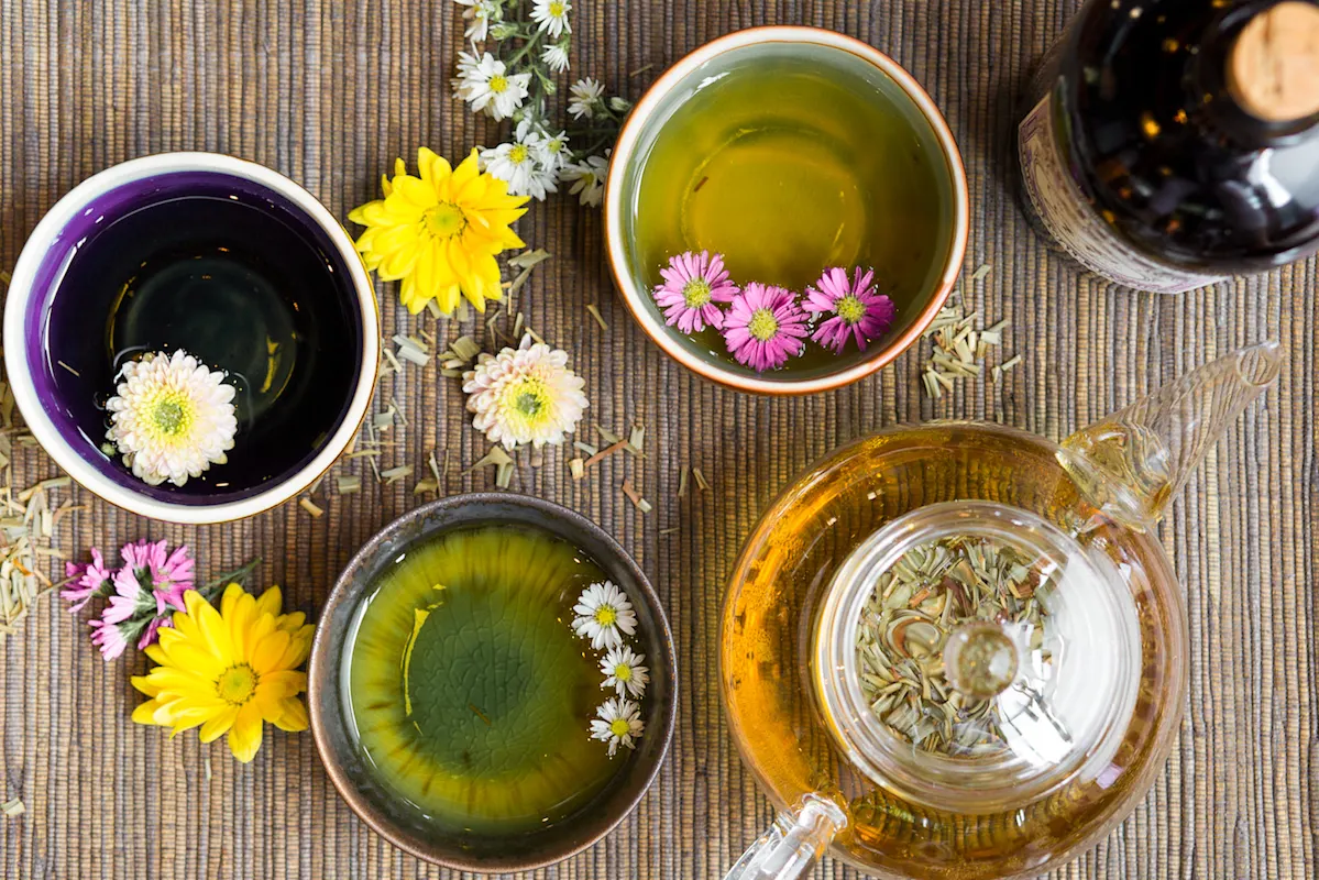 Kräutertee in vier Tassen mit Teekanne und Blumen auf gestreiftem Tisch.