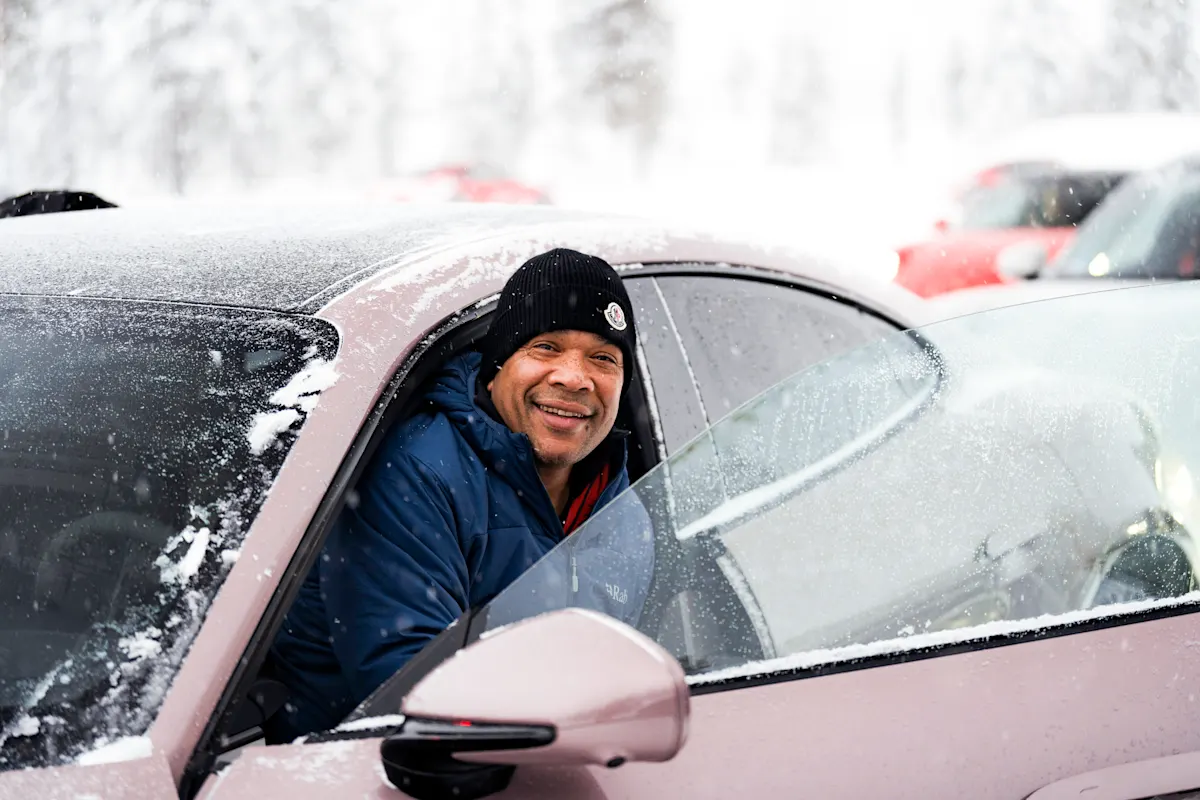 Smiling man in winter hat sits in snow-covered Porsche with open door.