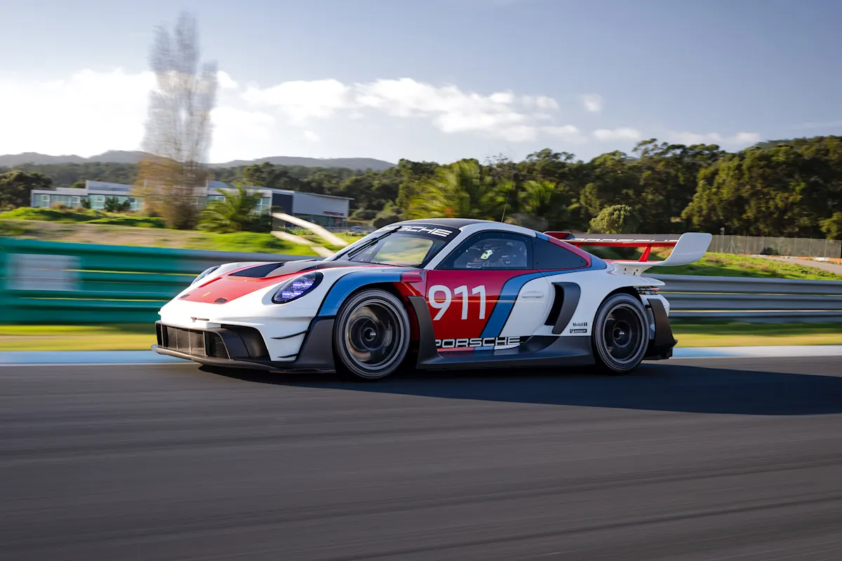 Porsche sports car speeds on track with scenic backdrop and clear blue sky.