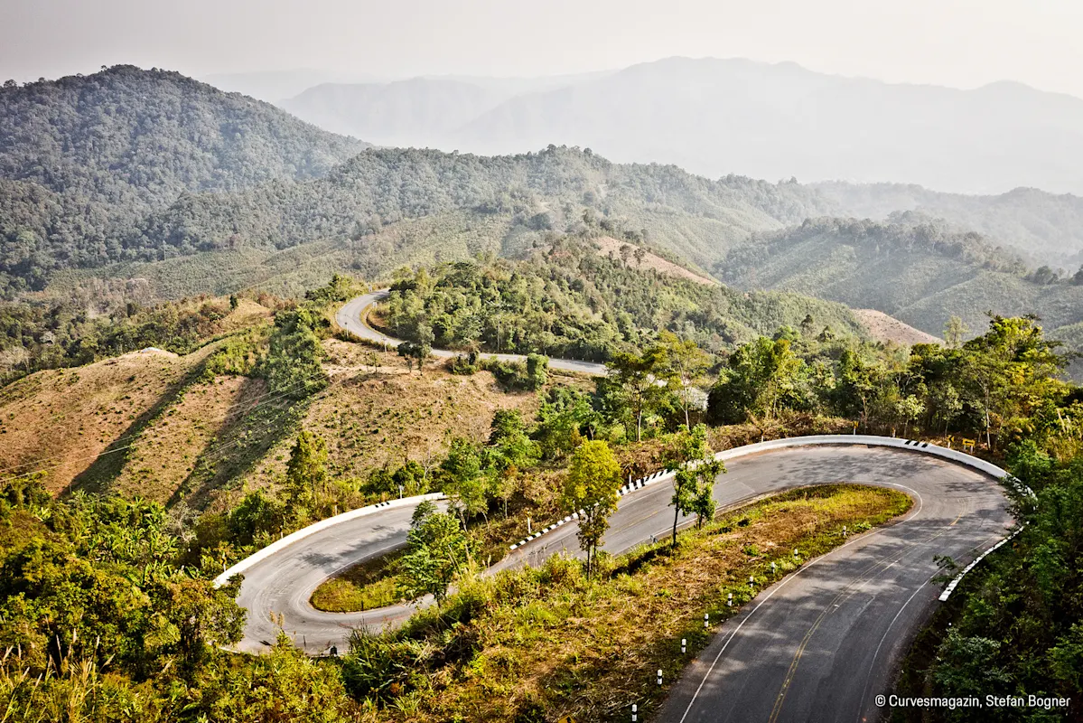 Kurvige Bergstraße zwischen grünen Hügeln und fernen Bergen.