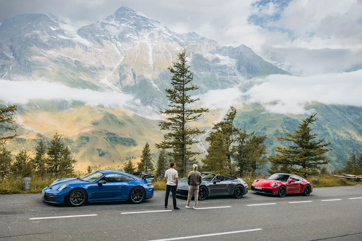 Zwei Personen neben drei Porsche Sportwagen auf malerischer Bergstraße.