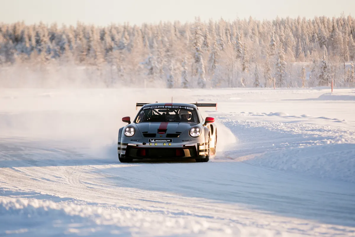 A Porsche race car drives on a snowy track with a snowy forest background.