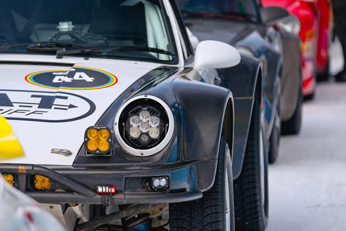 White Porsche race car with black hood and colorful decals parked in a row with other cars in background.