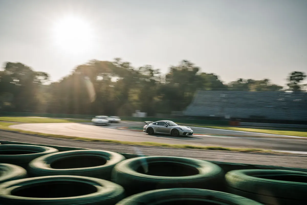 A silver Porsche 911 GT3 speeds along a race track in bright sunlight, followed by two other cars, with green tire barriers in the foreground.