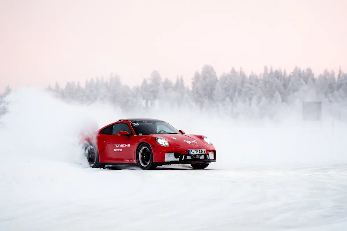 Red Porsche drifting on snowy road, snow spraying, trees in background.