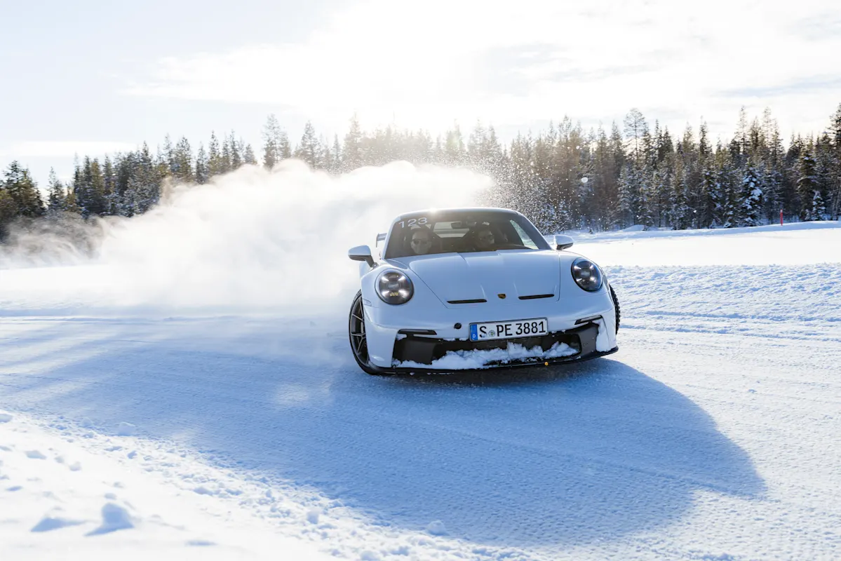 A white Porsche drifts on snowy track, kicking up snow with trees and bright sky.