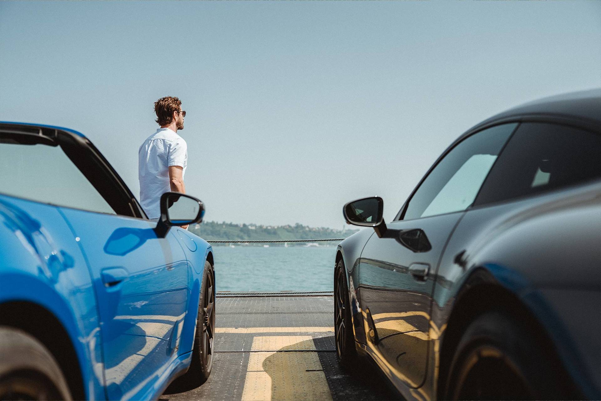 Mann steht zwischen blauem Cabrio und schwarzem Sportwagen am Wasser.