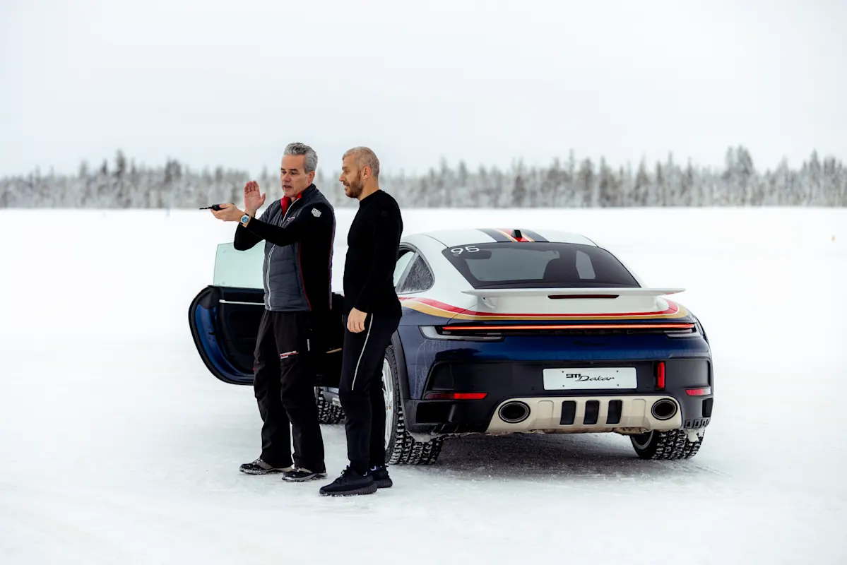 Two men beside a Porsche sports car on snowy terrain, one gesturing, the other observing.