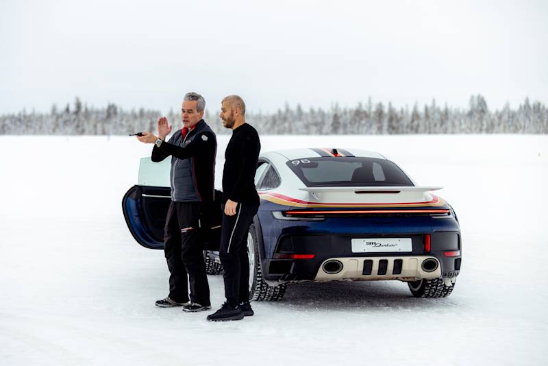 Two men beside a Porsche sports car on snowy terrain, one gesturing, the other observing.