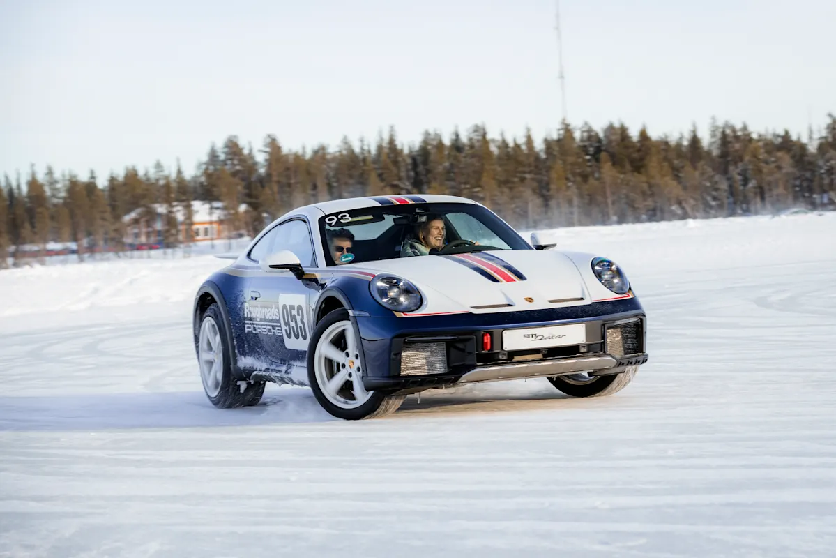 Porsche sports car drifts on snowy track with two people inside, pine trees in background.