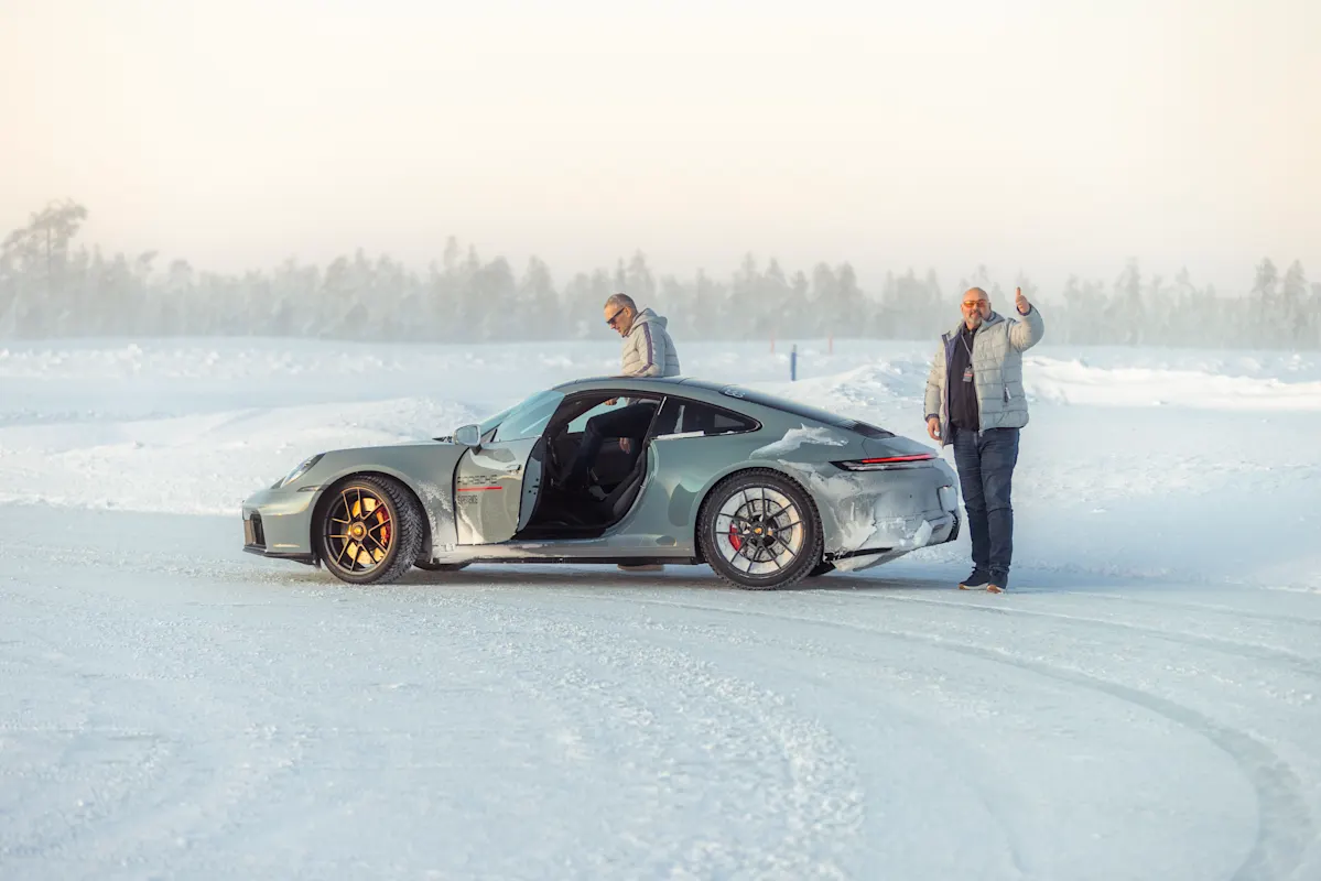 Two people with a silver Porsche on snowy landscape, one by the car and one behind it.