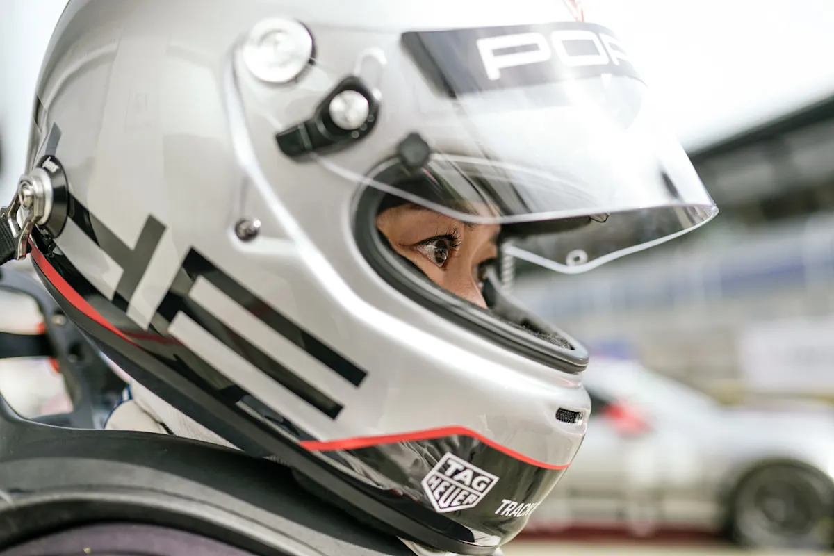 Person in racing gear, focused with visor up, cars blurred in background.