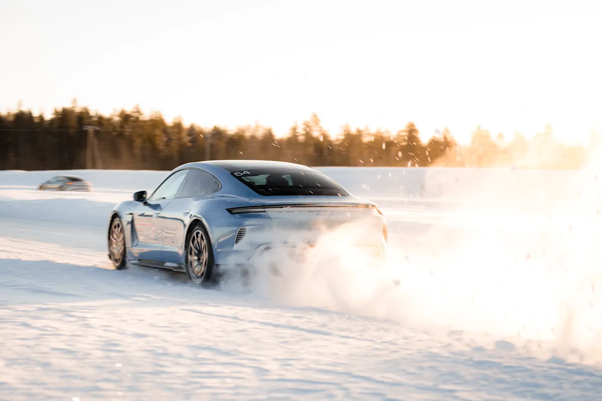 A silver Porsche speeds on snowy road, kicking up snow, with trees and another car in background.