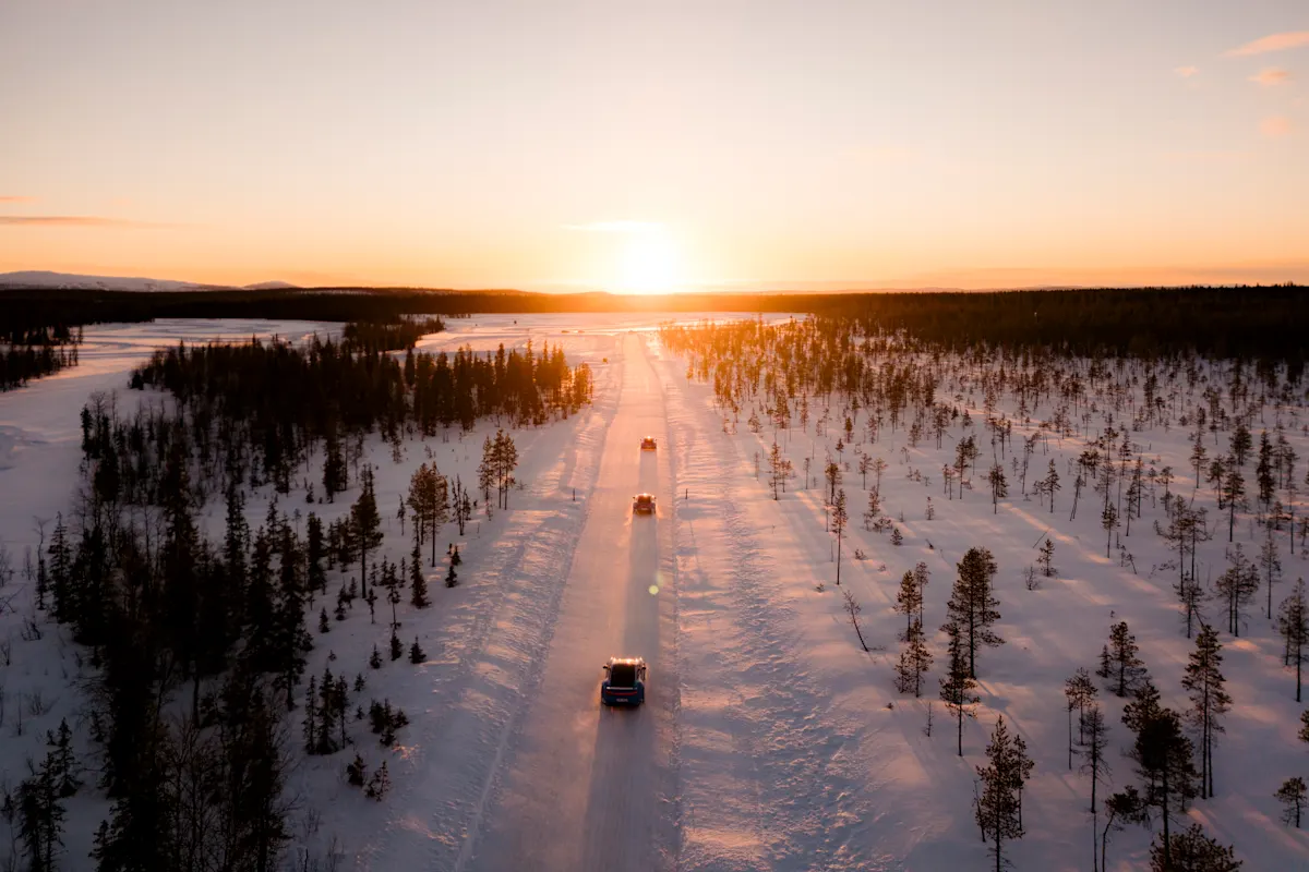 Convoy of cars on a snowy forest road at sunset, casting long shadows across the landscape.