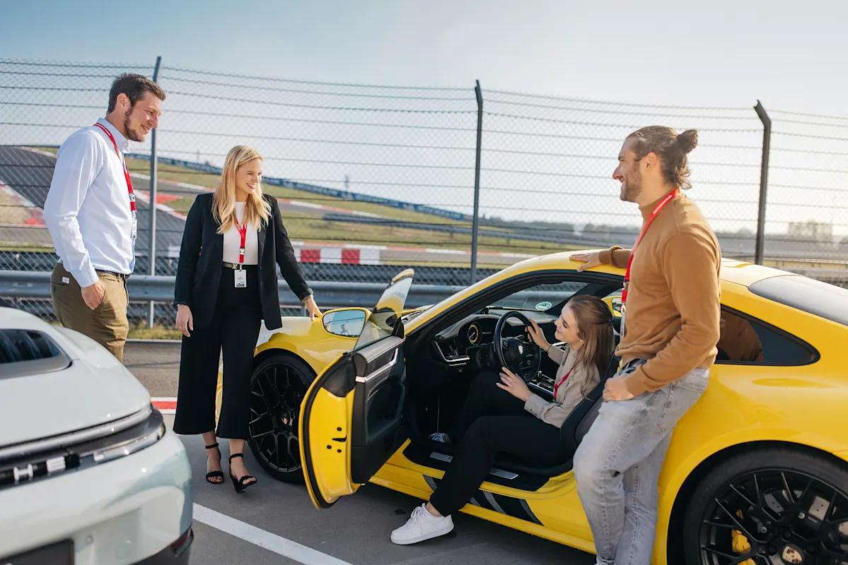 Four people at a Porsche on a test track, one woman seated at the wheel.