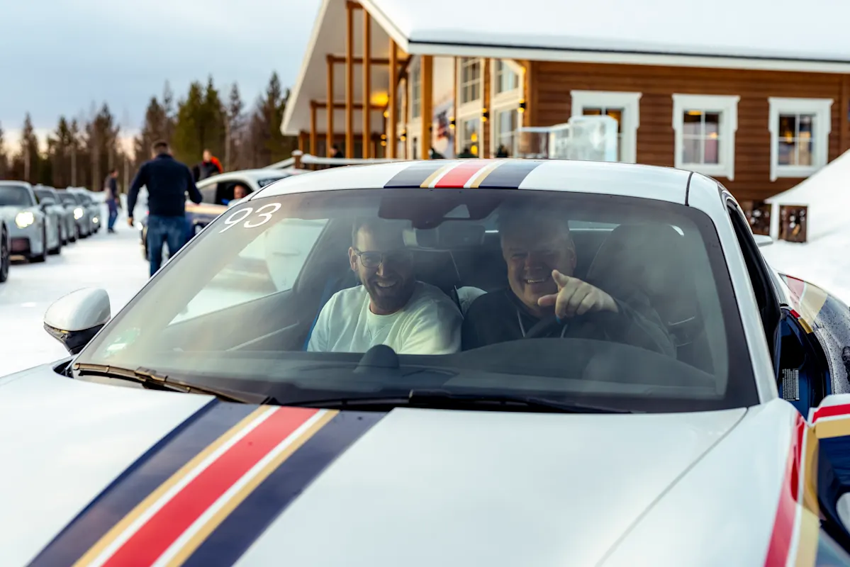 Two men smiling in a Porsche with racing stripes parked near a wooden building on a snowy day.
