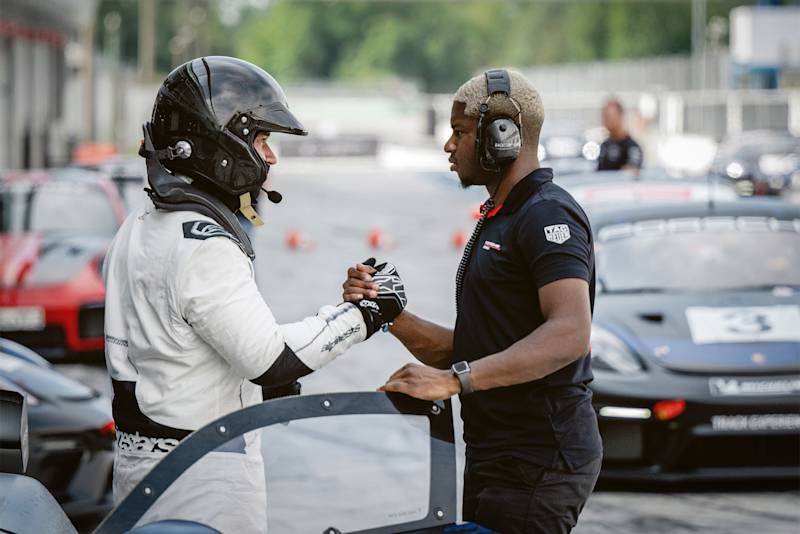 Ein Teilnehmer der Porsche Track Experience in Rennbekleidung gibt einem Instrukteur in der Boxengasse die Hand, umgeben von Porsche Sportwagen.