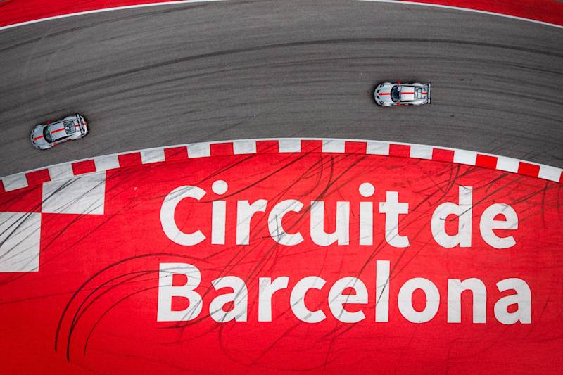 Luftaufnahme von zwei Porsche GT Fahrzeugen in einer Kurve des Circuit de Barcelona mit markanten roten Streckenmarkierungen und Reifenspuren.