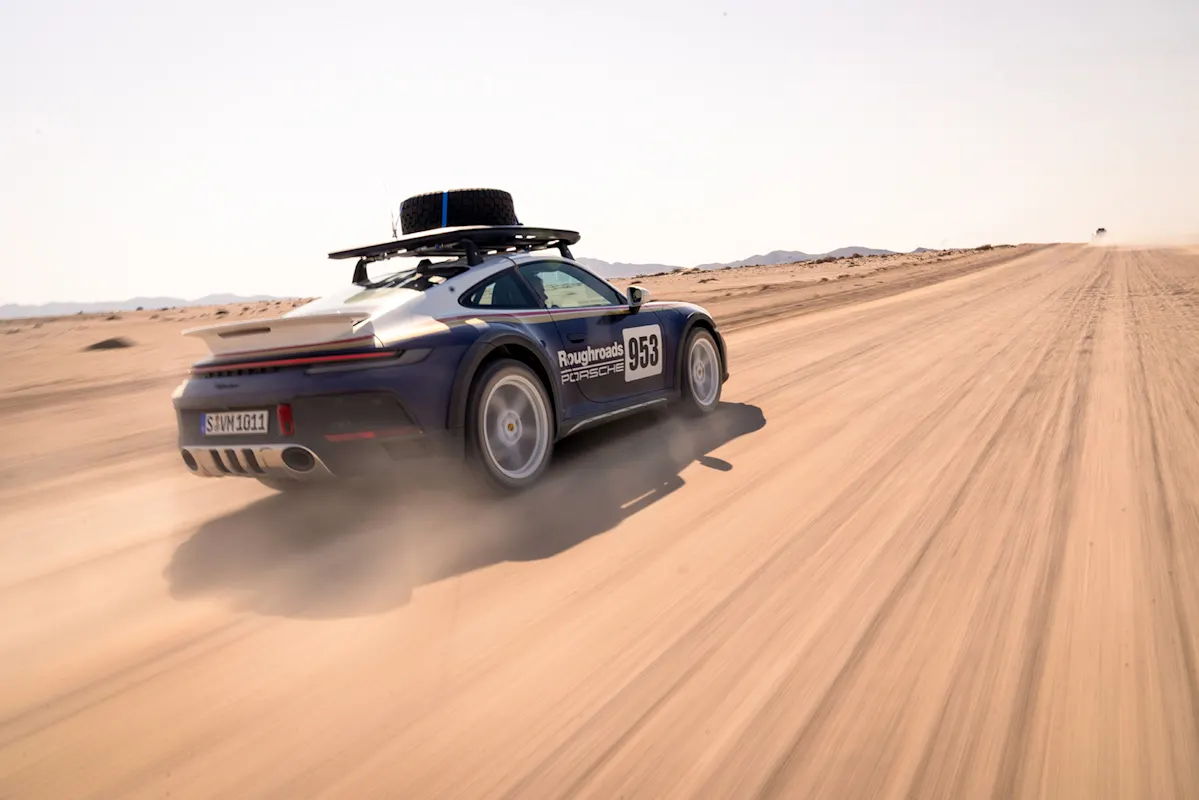 Porsche mit Dachträger rast auf Wüstenstraße, Staubwolke im klaren Himmel.