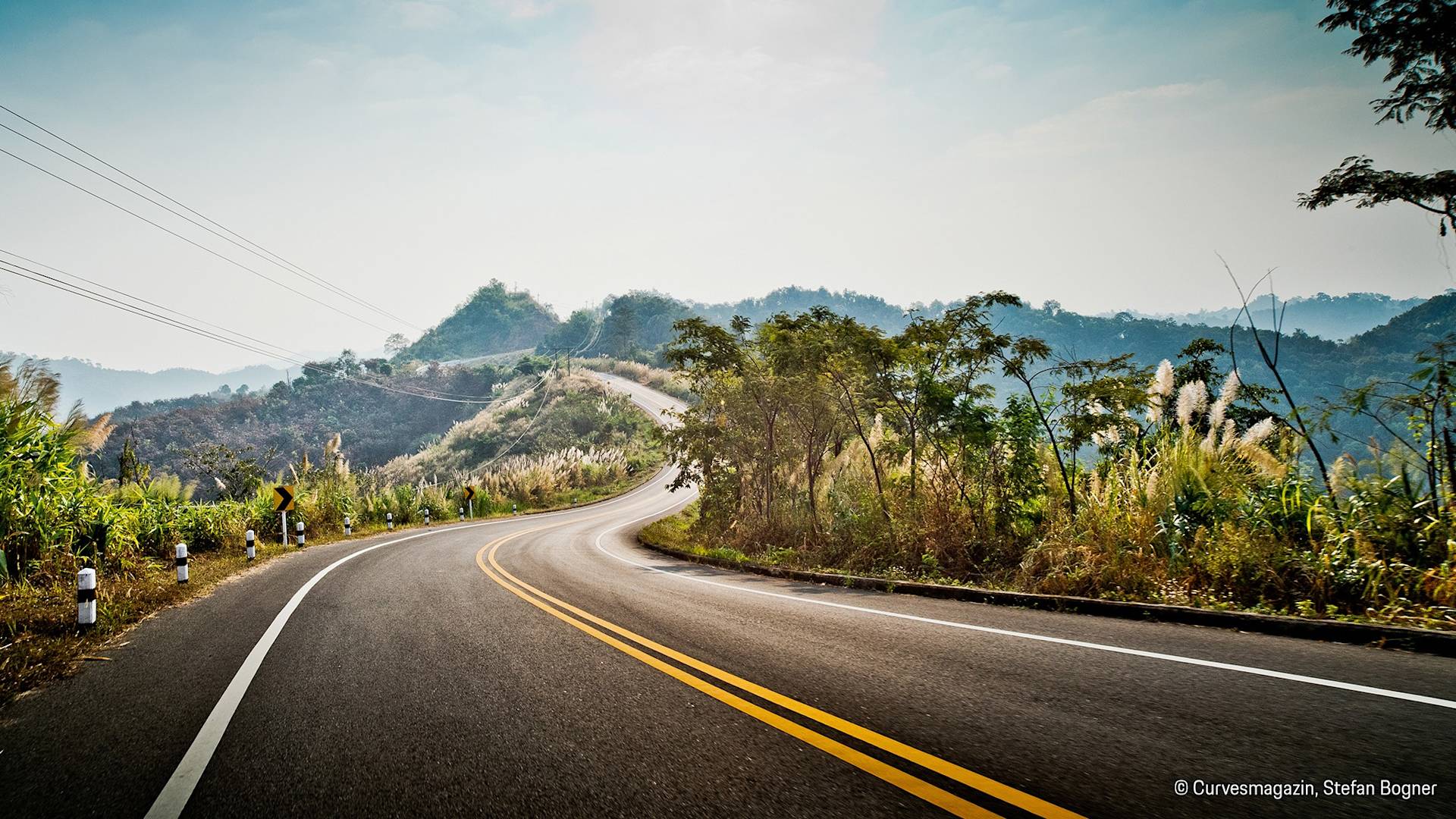 Geschwungene Straße durch grüne Hügel unter teilweise bewölktem Himmel.