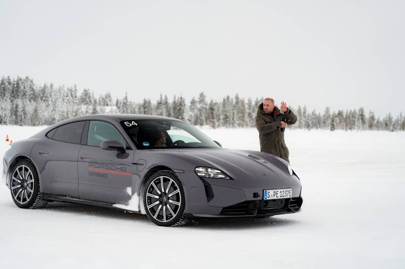 Instructor giving instructions next to a Slate Grey Neo coloured Porsche on a snowy surface with forest in the background.