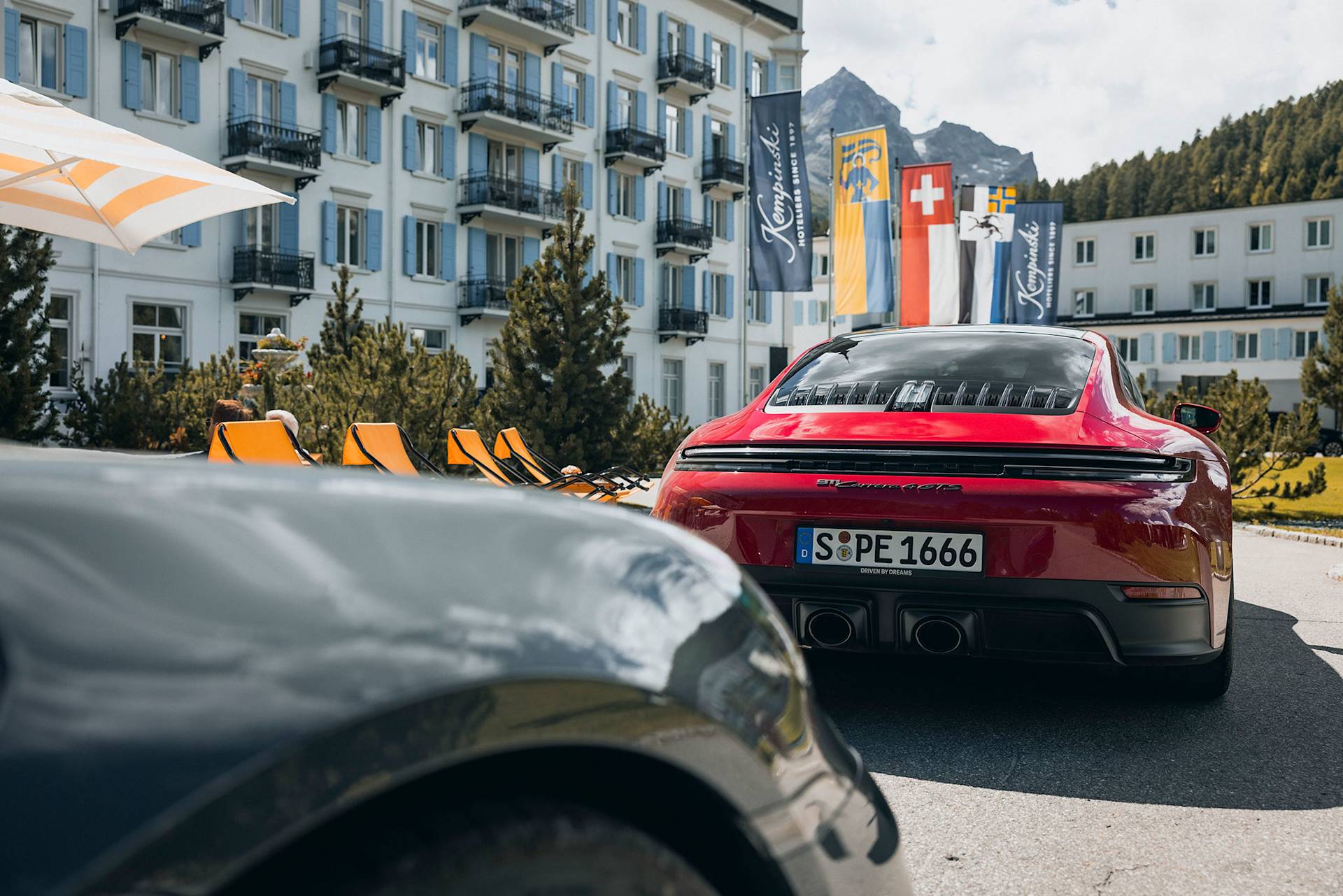 Red Porsche Taycan in front of a hotel with flags and mountains.