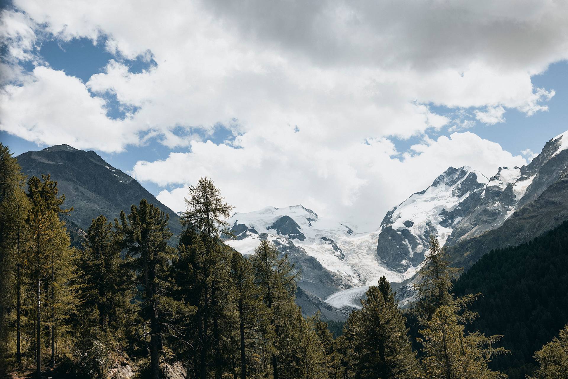 Mountains with snow, pines, and clouds on a sunny day.