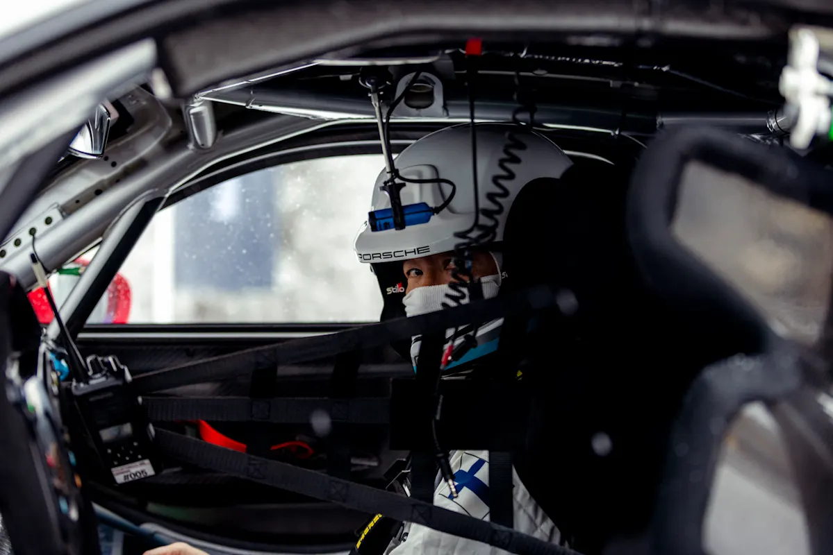 Close-up of a racing driver in full gear inside a Porsche cockpit, focused before a snowy track session.