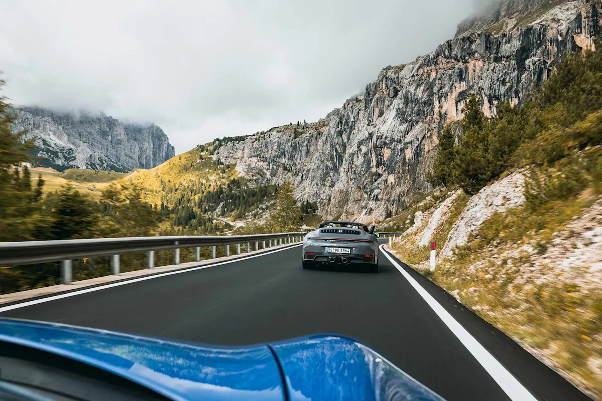 Zwei Porsche Sportwagen auf Gebirgsstraße mit Klippen und Grün.
