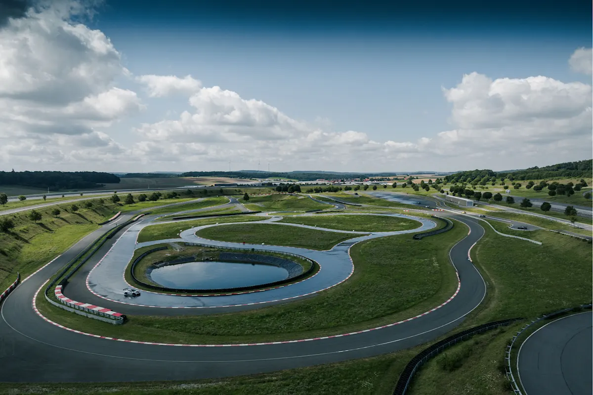 Aerial view of a winding Porsche racetrack with a pond, greenery, and a cloudy blue sky.