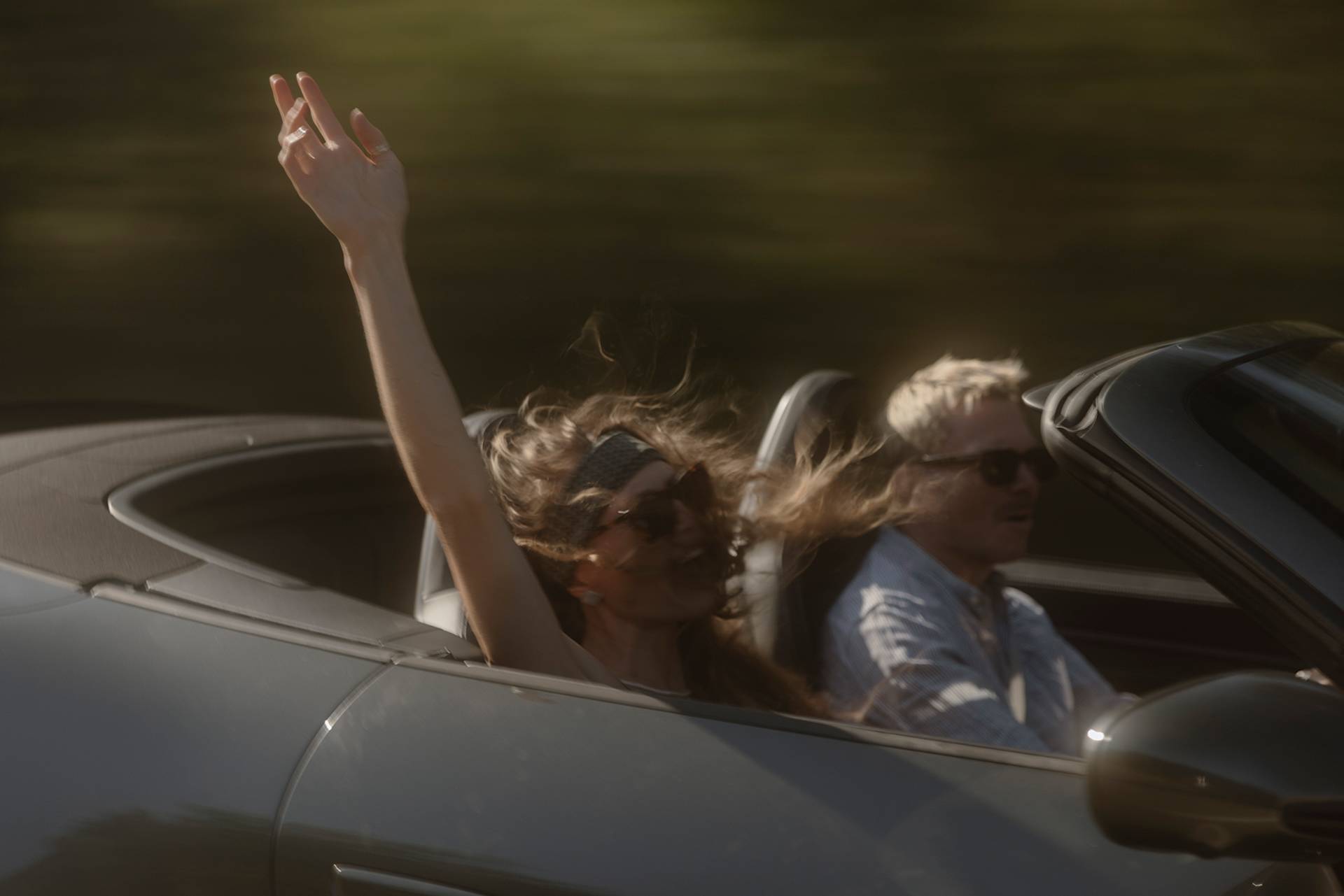 Zwei Passagiere in einem Cabrio, einer hebt freudig den Arm.