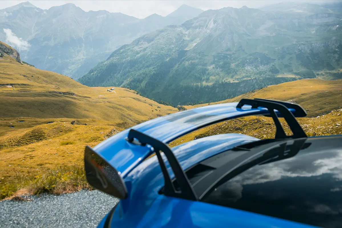 Blauer Porsche auf Bergstraße, grüne Hügel und Berge in der Ferne.