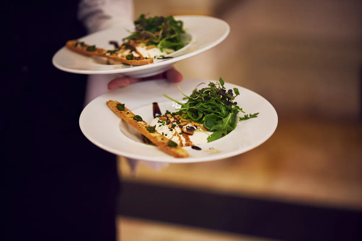 Kellner mit Tellern voll Salat, Brotstangen, Käse und Balsamico-Glasur.