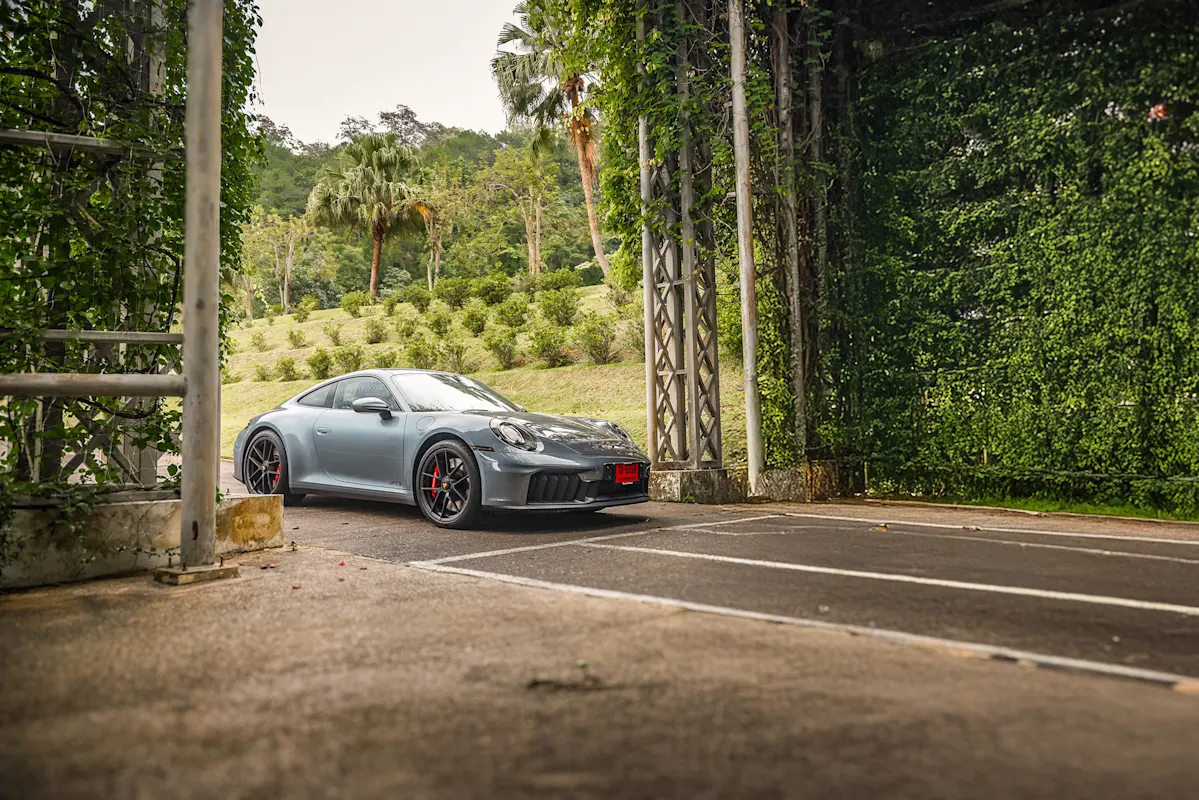 Grauer Porsche steht neben einem Garten mit Bäumen und Hecken.