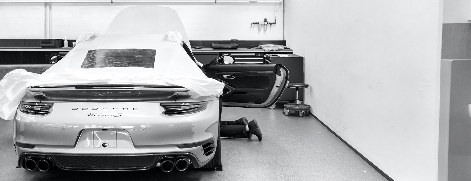 Ein Schwarz-Weiß-Foto eines Porsche 911 Turbo S mit sichtbarem Heck und einem Mechaniker, der unter der geöffneten Motorhaube in einer Werkstatt arbeitet.