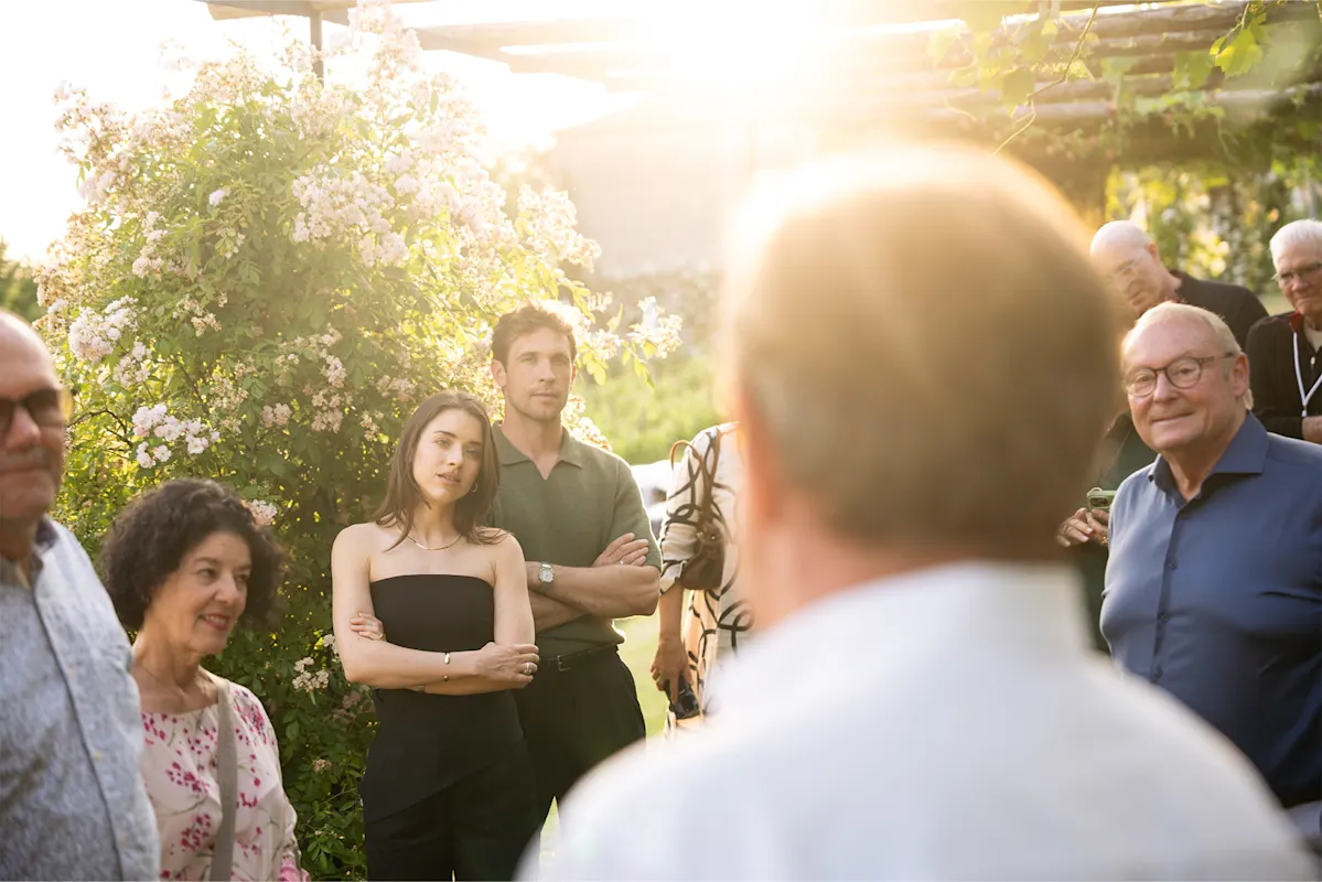 Menschenmenge hört Redner im Freien zu, umgeben von Grün und Blumen.