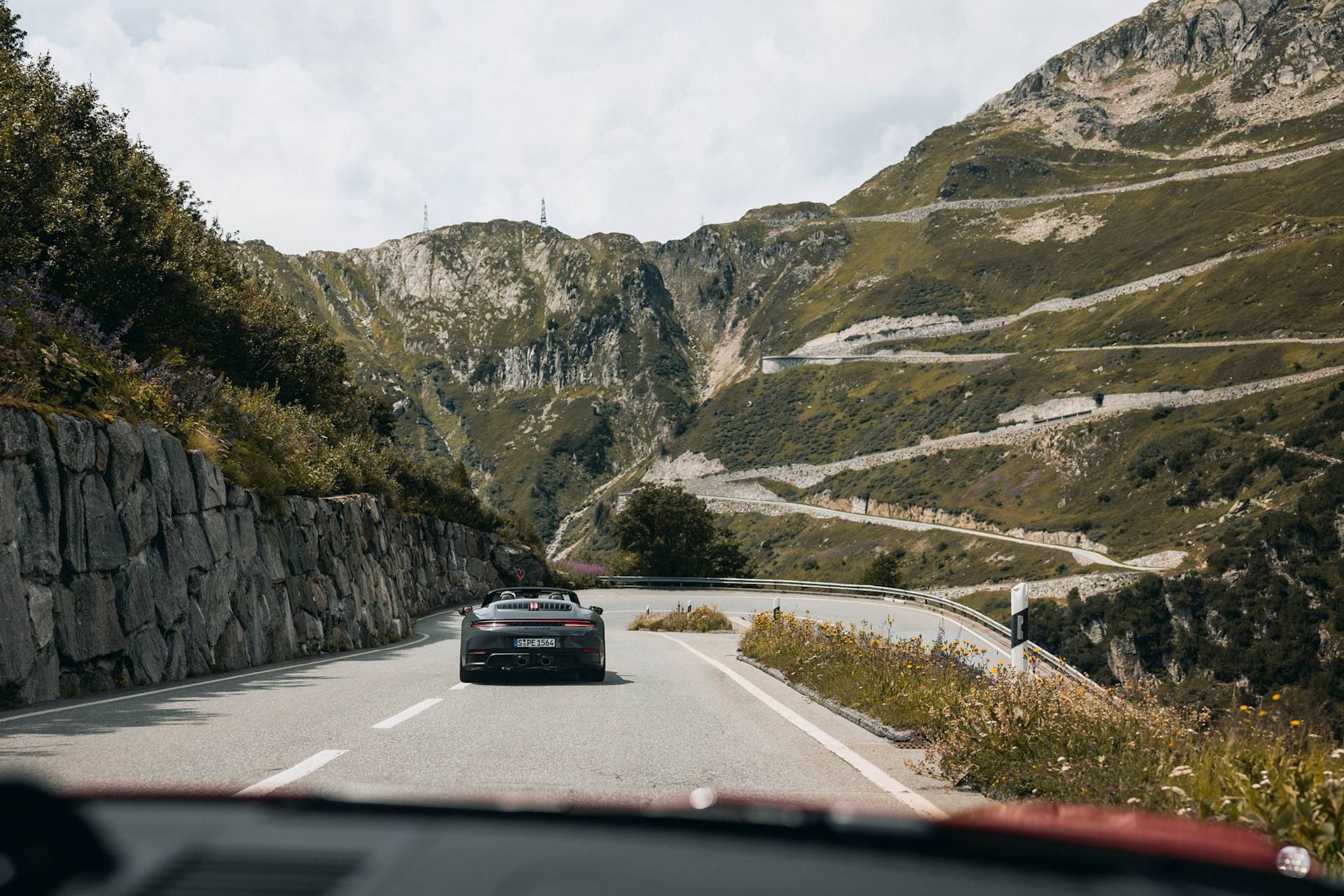 Black convertible Porsche on a mountain road with greenery and clouds.