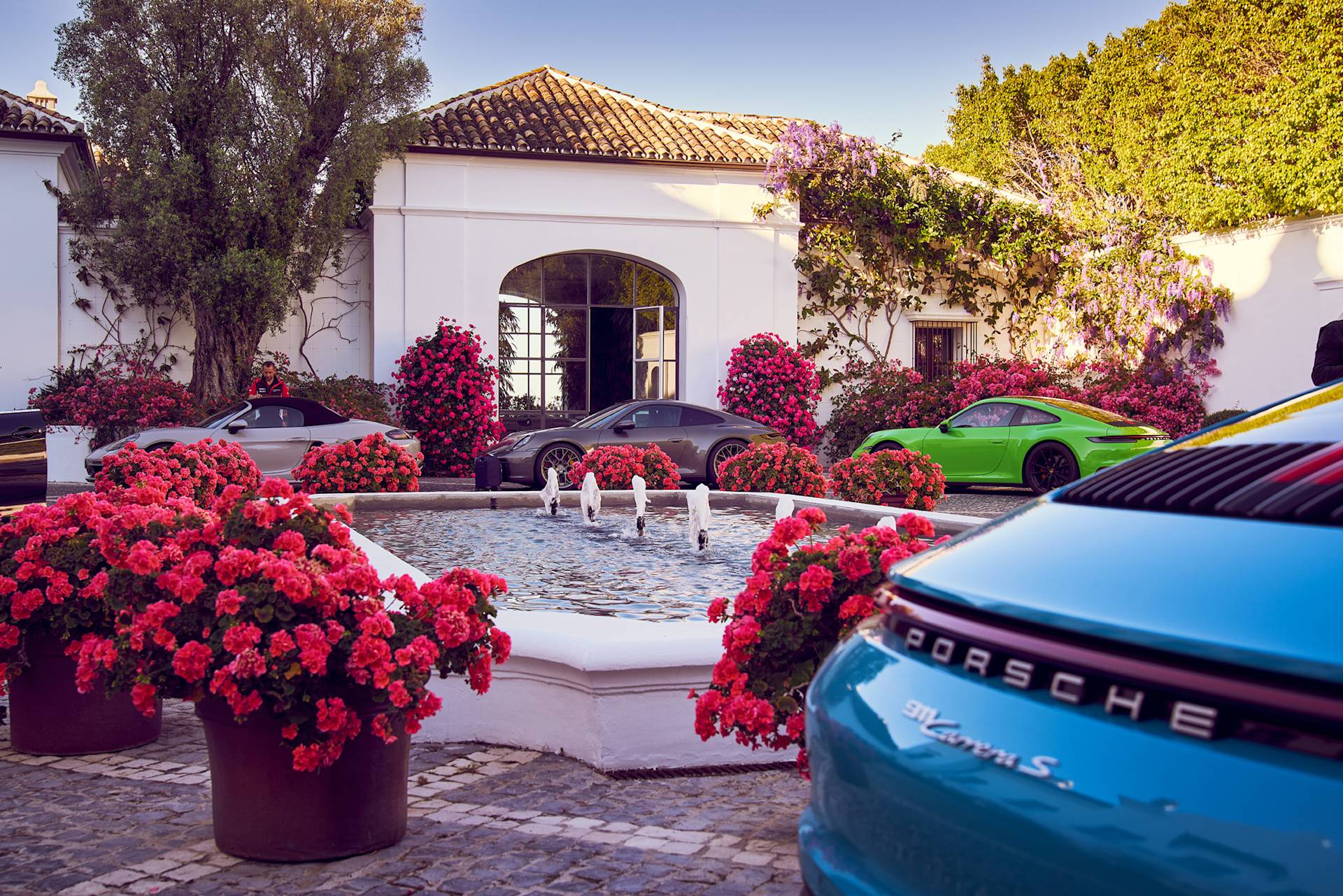 Innenhof mit rosa Blüten, Brunnen und buntem Porsche parkend.
