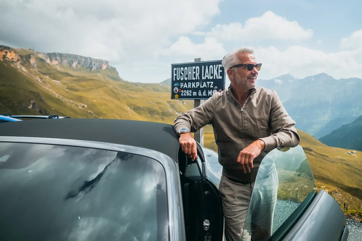 Mann mit Sonnenbrille neben Cabrio-Porsche bei Fuscher-Lacke-Schild und Bergen.