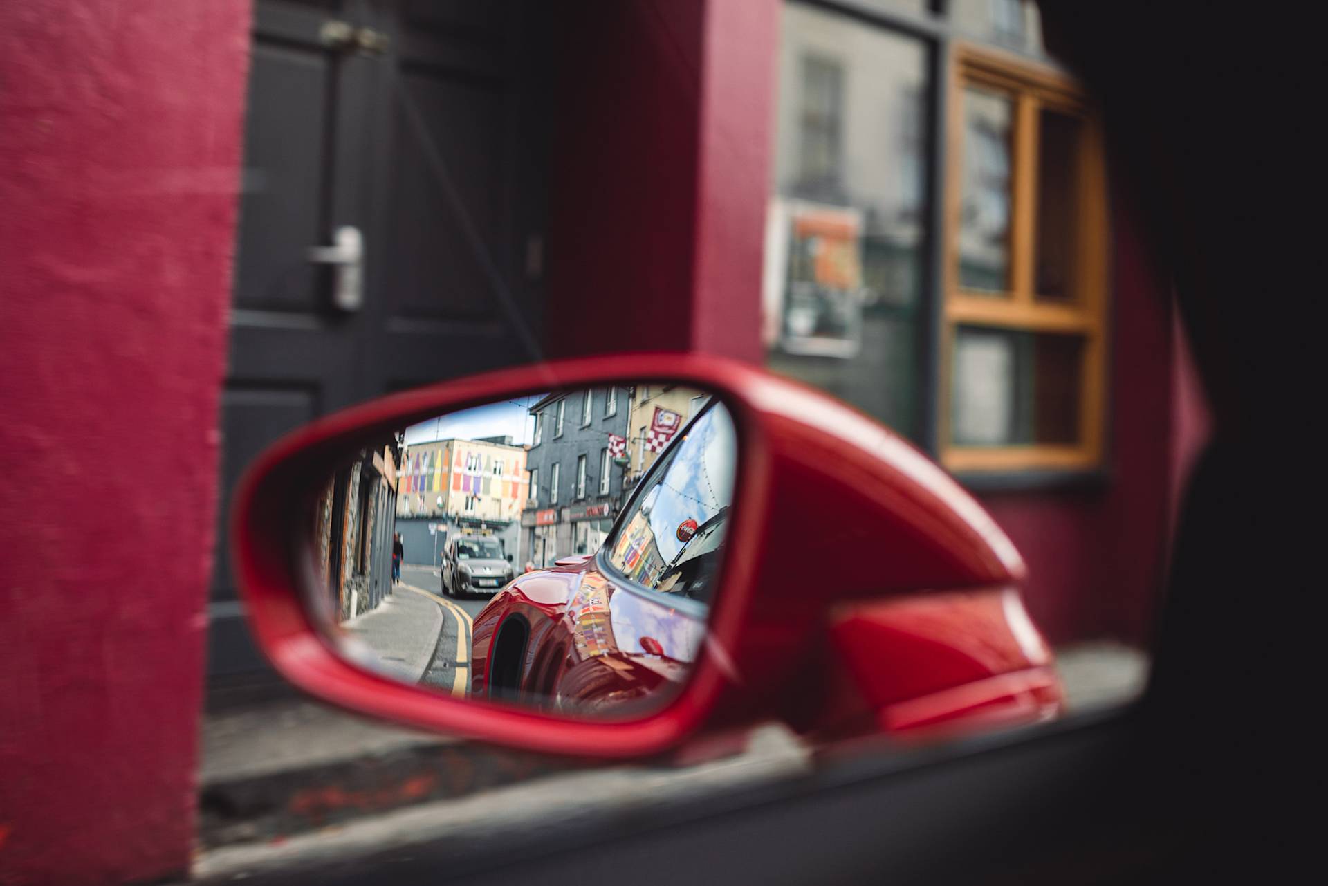 Seitenspiegel eines roten Autos spiegelt lebhafte Straßenszene.