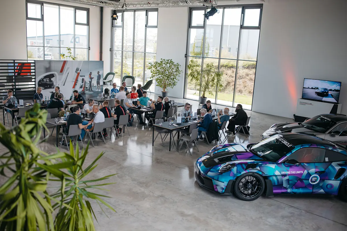 People in a modern room with large windows; colorful Porsche sports cars displayed nearby.