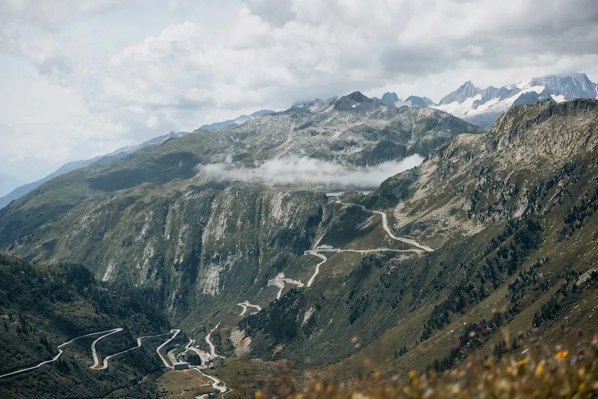 Mountain road through green hills and rocky peaks under clouds.