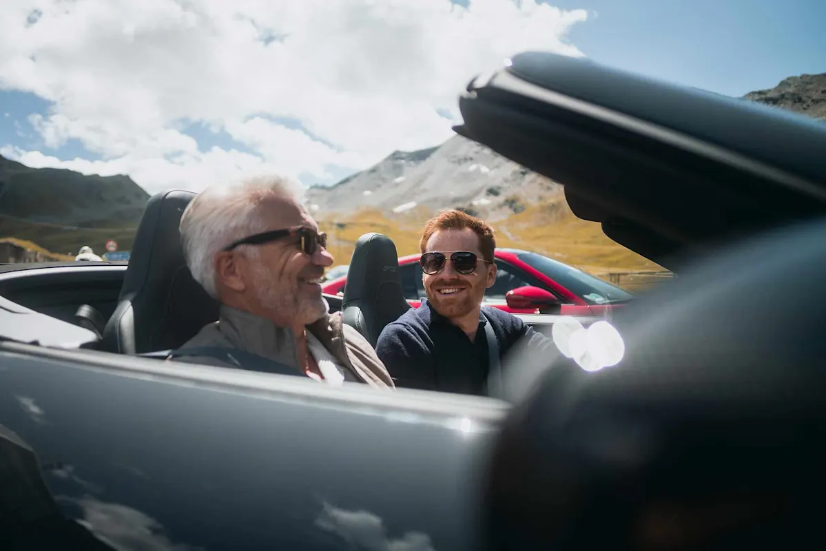 Zwei Männer lächeln in einem Cabrio vor Bergen und blauem Himmel.