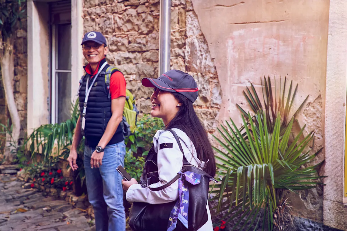Zwei Personen in Hüten lächeln an einer Steinmauer mit Grün.
