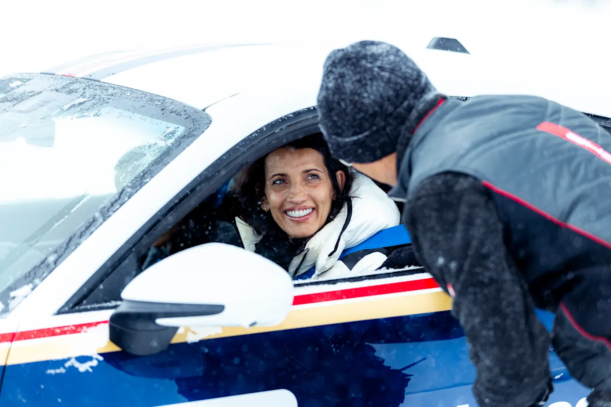 Woman smiling in Porsche talks to man leaning in through window on snowy day.