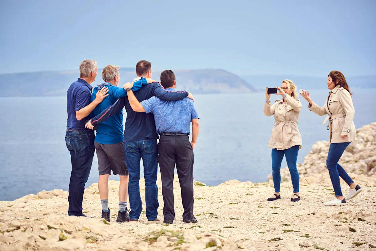 Vier Männer am Meer, zwei Frauen fotografieren sie an felsiger Küste.