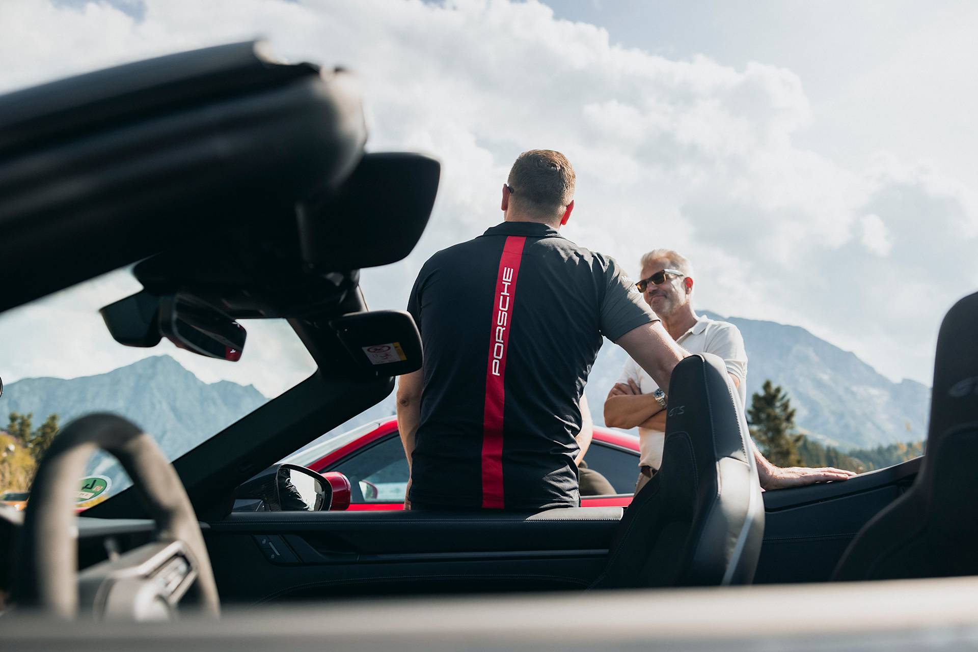 Zwei Männer plaudern an einem sonnigen Tag bei einem Cabrio-Porsche nahe Bergen.
