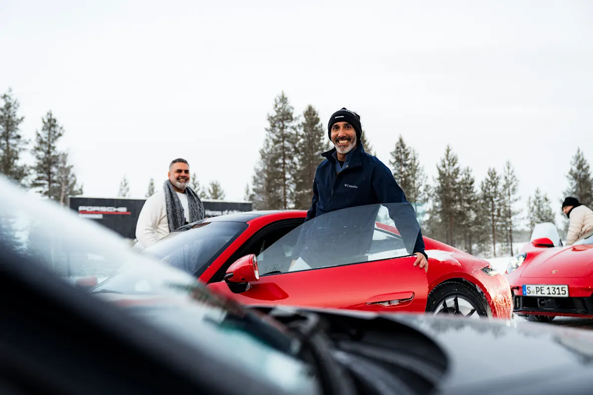 Two men smiling while getting into a red Porsche, winter setting with pine trees in the background.