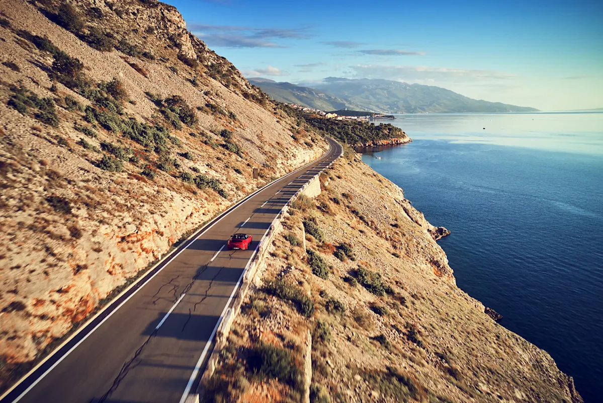 Roter Porsche an Küstenstraße bei Klippe und Meer unter klarem Himmel.