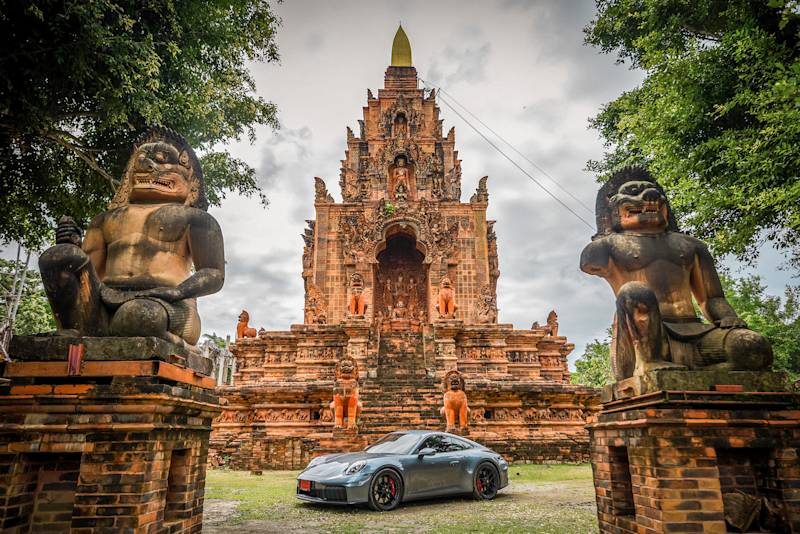 Silberner Porsche vor Tempel mit Statuen und Bäumen.